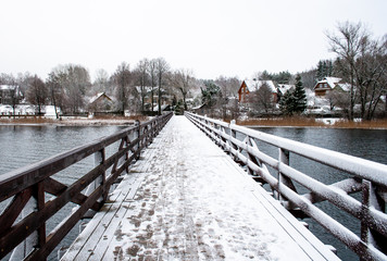 Wooden bridge in winter, trees and and wooden houses covered by snow in a village