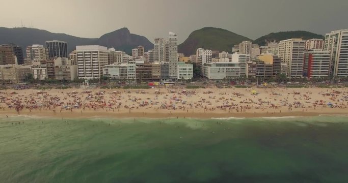 Aerial tracking shot along Ipanema beach with sea in foreground and hotels apartment blocks and mountains in background