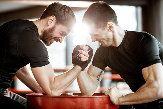 Two young athletes in black sportswear having a hard arm wrestling competition on a red barrel in the gym