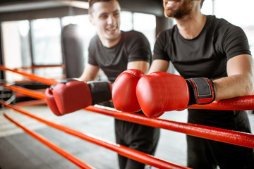 Two athletic men in black sportswear talking together, having fun during the break on the boxing ring at the gym. Image focused on the gloves