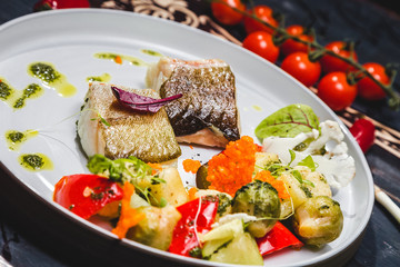 fish with steamed vegetables on a white plate. wooden background decorated with vegetables