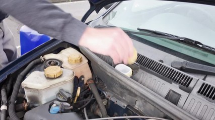 A caucasian, middle-aged, British man topping up the screenwash fluid in his small petrol car.