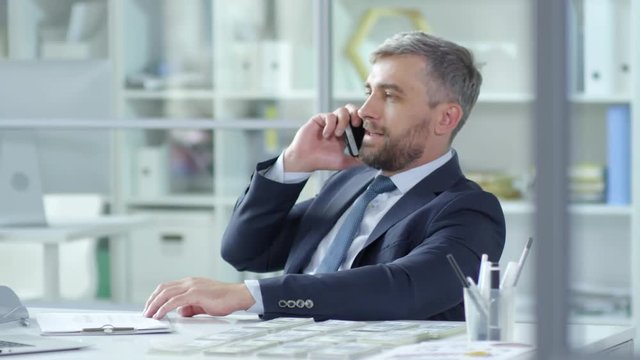 Tracking shot of mid-aged bearded businessman in formal suit talking on mobile phone while sitting at desk with money stacks on it in the office
