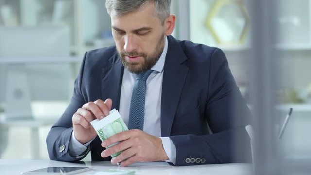 Experienced Caucasian businessman counting banknotes and typing on digital tablet while working at desk in the office - Powered by Adobe