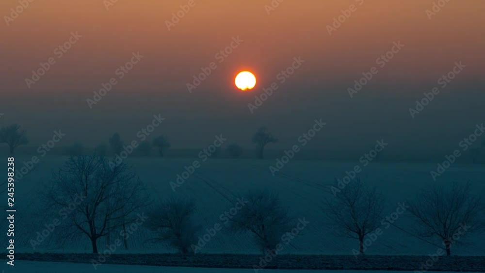 Poster sunset time lapse on a snowy landscape with fields and trees, camera is zoomed and follows the sun