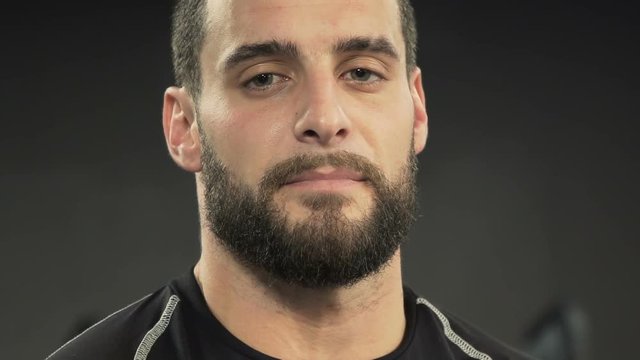 Close up shot of tired athletic handsome young man with beard in black sportswear looking at camera, changing body position. Fatigue concept