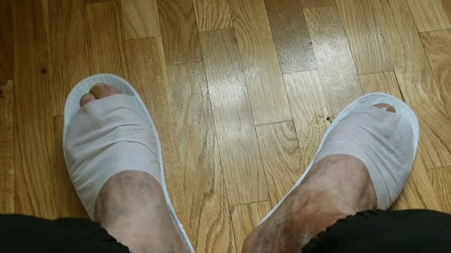 Person bangs his feet, which have disposable slippers at wooden floor. Fragment feets close-up. High angle view.