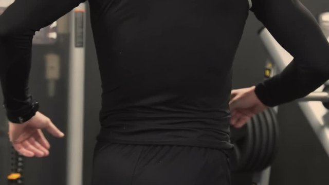 Side view of athletic handsome young man with beard in black sportswear stretching back muscles after exercising in gym, pressing hands against lower back, bending. Health concept