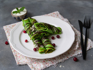 Mint thin crepes of green color, twisted into tubes, with chocolate sauce, sour Cream, mint leaf and almond petals on a white plate on gray background with floral fabric. Breakfast. Selective focus.