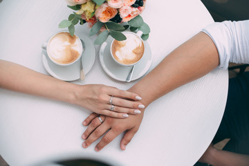 The bride and groom hold hands, holding expensive wedding rings with white gold, on the background of a wedding bouquet and two cups of coffee on a white table.