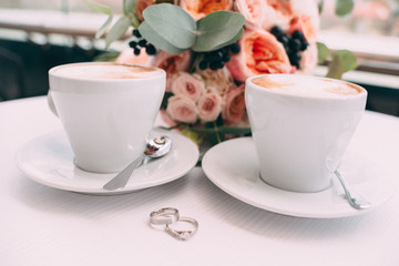 wedding rings on a white table on the background of a wedding bouquet and two cups of coffee.
