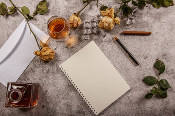 Mock up background with men's hand holding a glass of whiskey, brandy withered flowers of white roses with a clean envelope, notepad and cup of coffee on a gray concrete background. View from above. 