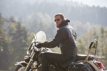 Back view of bearded motorcyclist with long hair in black leather jacket sitting on cruiser...