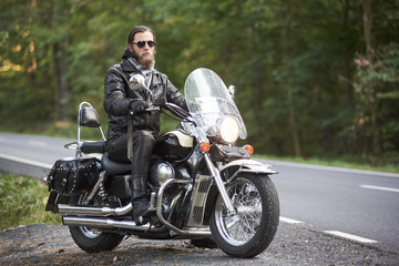 Fototapeta na wymiar Handsome bearded motorcyclist in black leather clothing and dark sunglasses riding on motorcycle on country roadside on background of empty straight asphalt road and green trees bokeh foliage.
