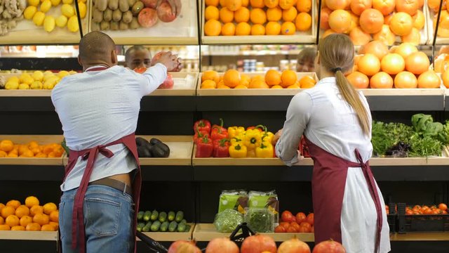 Multi ethnic couple of small business owners in aprons arranging groceries on stand in grocery store. Positive diverse entrepreneurs busy filling up fruit and vegetable storage racks at minimarket.
