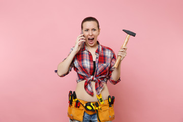 Angry handyman woman in plaid shirt kit tools belt full of instruments talking on mobile phone, conducting unpleasant conversation isolated on pink background. Female in male work. Renovation concept