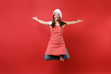 Housewife female chef cook or baker in striped apron white t-shirt, toque chefs hat isolated on red wall background. Full length portrait housekeeper woman jumping high up. Mock up copy space concept.