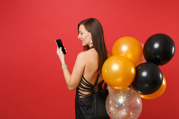 Back rear view of young woman in black dress holding, using mobile phone with blank black empty screen, air balloons isolated on red background. Happy New Year, birthday mockup holiday party concept.