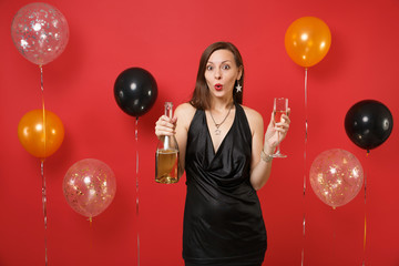 Amazed young girl in black dress celebrating holding glass, bottle of champagne on bright red background air balloons. International Women's Day, Happy New Year, birthday mockup holiday party concept.