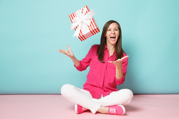 Full length portrait woman in rose shirt blouse white pants sitting on floor hold present box...