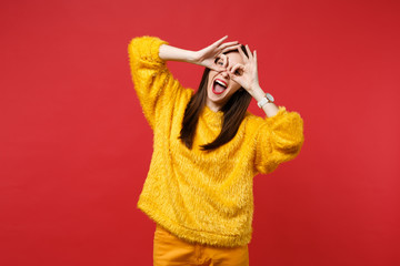 Funny young woman in yellow fur sweater holding hands near eyes, imitating glasses or binoculars isolated on bright red wall background. People sincere emotions, lifestyle concept. Mock up copy space.