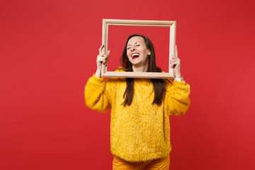 Portrait of laughing young woman in yellow fur sweater with closed eyes holding picture frame isolated on red wall background in studio. People sincere emotions, lifestyle concept. Mock up copy space.