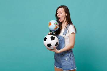 Shocked young woman football fan holding soccer ball, looking on Earth world globe isolated on blue turquoise background. People emotions, sport family leisure lifestyle concept. Mock up copy space.