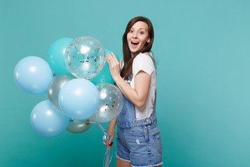 Fototapeta premium Surprised young woman in denim clothes keeping mouth wide open, celebrating, holding colorful air balloons isolated on blue turquoise wall background. Birthday holiday party, people emotions concept.