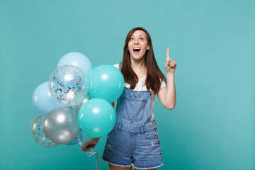 Fototapeta premium Surprised young woman in denim clothes looking, pointing index finger up, celebrating hold colorful air balloons isolated on blue turquoise background. Birthday holiday party, people emotions concept.