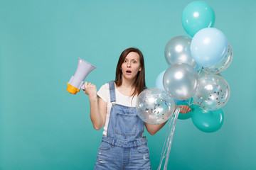 Shocked perplexed young woman keeping mouth open holding megaphone, colorful air balloons while celebrating isolated on blue turquoise wall background. Birthday holiday party, people emotions concept.