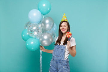 Smiling young woman in denim clothes, birthday hat holding round lollipop celebrating with colorful...