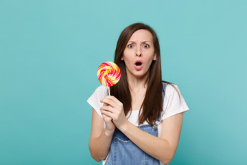 Portrait of shocked young woman in denim clothes keeping mouth wide open, holding colorful round...