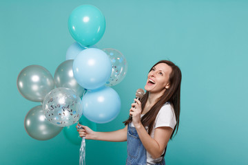Fototapeta premium Charming young woman in denim clothes looking up sing song in microphone, celebrating hold colorful air balloons isolated on blue turquoise background. Birthday holiday party, people emotions concept.