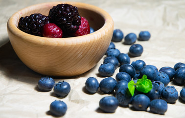 An isolated wooden bowl full of blueberries and wildberries with plain soft background decorated by fresh mint and with a few blueberries around on the paper