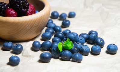 An isolated wooden bowl full of blueberries and wildberries with plain soft background decorated by fresh mint and with a few blueberries around on the paper
