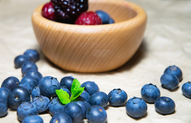 An isolated wooden bowl full of blueberries and wildberries with plain soft background decorated by fresh mint and with a few blueberries around on the paper