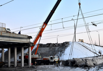 Truck crane, concrete slab hanging from crane hook above building skeleton at construction site, construction of an automobile bridge