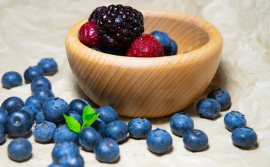 An isolated wooden bowl full of blueberries and wildberries with plain soft background decorated by fresh mint and with a few blueberries around on the paper