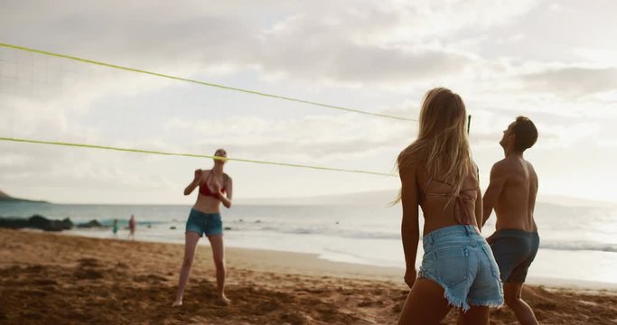 Active friends playing beach volleyball at sunset, athletic man spikes volleyball