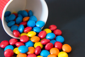 multicolored round dragee candies poured from an inverted paper cup on a black background