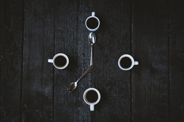 Coffee cups aranged as a clock face on a wooden black background. Four espresso mugs like a symbol of time with two spoons. Time for coffee. Breakfast time. Good morning concept.