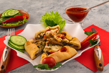 Mini pizza with cheese, mushrooms and slices of fresh vegetables. Spicy tomato sauce. Cutlery on a red napkin.