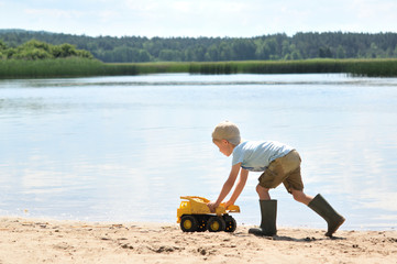 Five years old boy playing with a small truck on a summer lake beach.