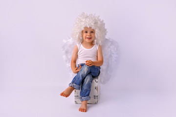 Angel boy, beautiful face smiling, in a white wig and angel wings. Dressed in white tank top and jeans, sitting on a crate in the studio on a white background. Children's innocence.