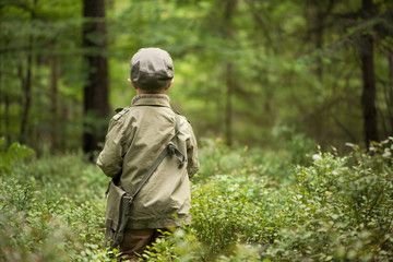 A boy in the woods, standing among trees, stands with his back turned. He is dressed in a green...