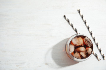 Glass of refreshing cola with ice cubes on white wooden background, top view. Space for text