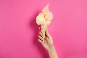 Woman holding ice cream cone with fluffy cotton candy on color background, closeup
