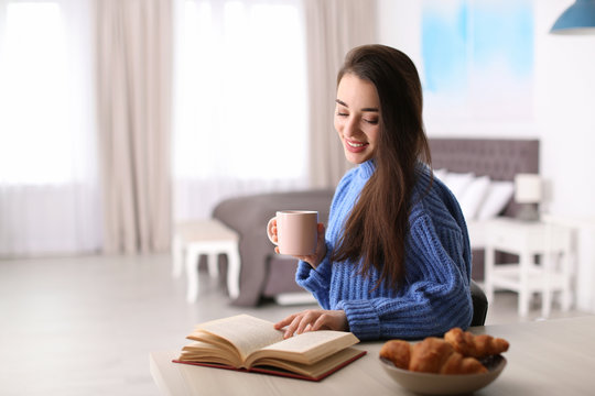 Young woman drinking coffee and reading book at table indoors, space for text. Winter season