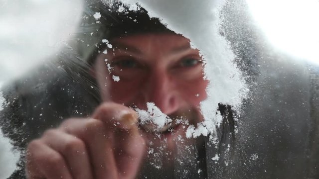 A man wipes the snow and draws on the frozen glass in the winter. A close-up portrait of a male who looks through a window from the yard.
