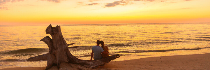 Romantic beach couple honeymoon banner background - Lovers enjoying watching sunset on summer travel destination sitting on tree trunk by the ocean.